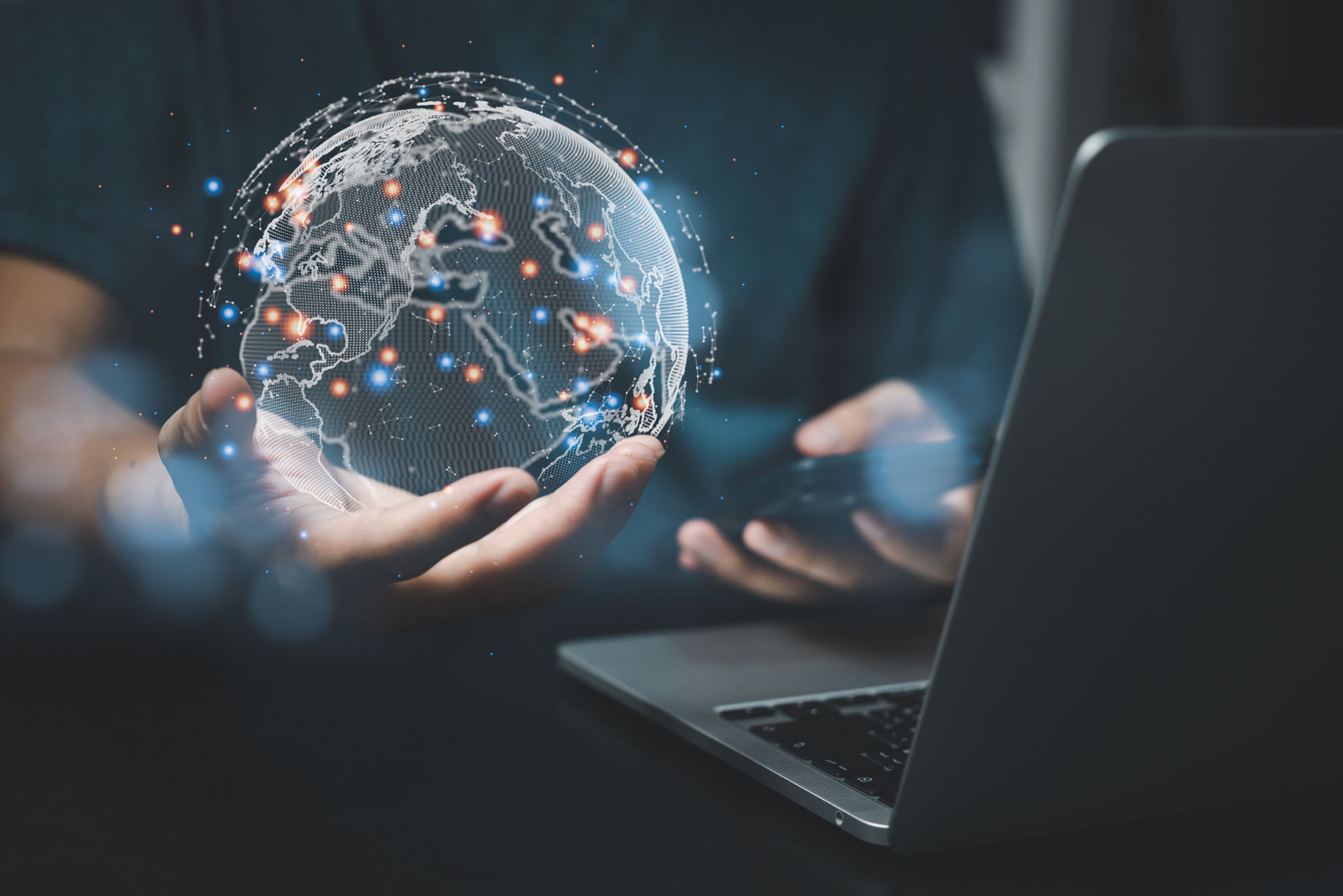 A man in a suit sitting infront of a laptop, holding out his hand with a 3D display of the globe in his hand